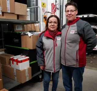 Zwei GO! Kurierinnen posieren nebeneinander im Lager. Im Hintergrund sind Pakete