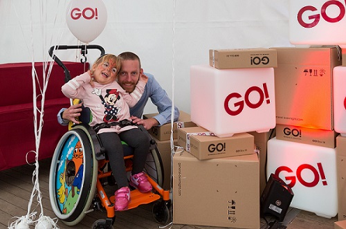 GO! Mitarbeiter F. Runzheimer hockt im Zimmer hinter einem Mädchen, auf einem Rollstuhl und umarmt diese. Das kleine Mädchen lacht vor Freude. Neben Ihnen viele GO! Pakete und ein Luftballon mit GO! Logo