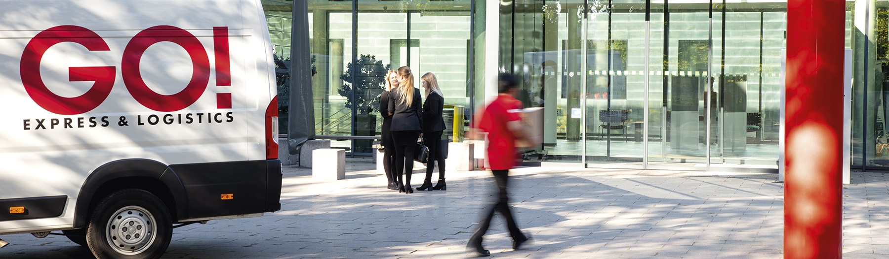 GO! Kurier läuft neben einem GO! Transporter auf die Eingangstüren des Forschungszentrum caesar in Bonn zu und im Hintergrund stehen drei Frauen in Businesskleidung