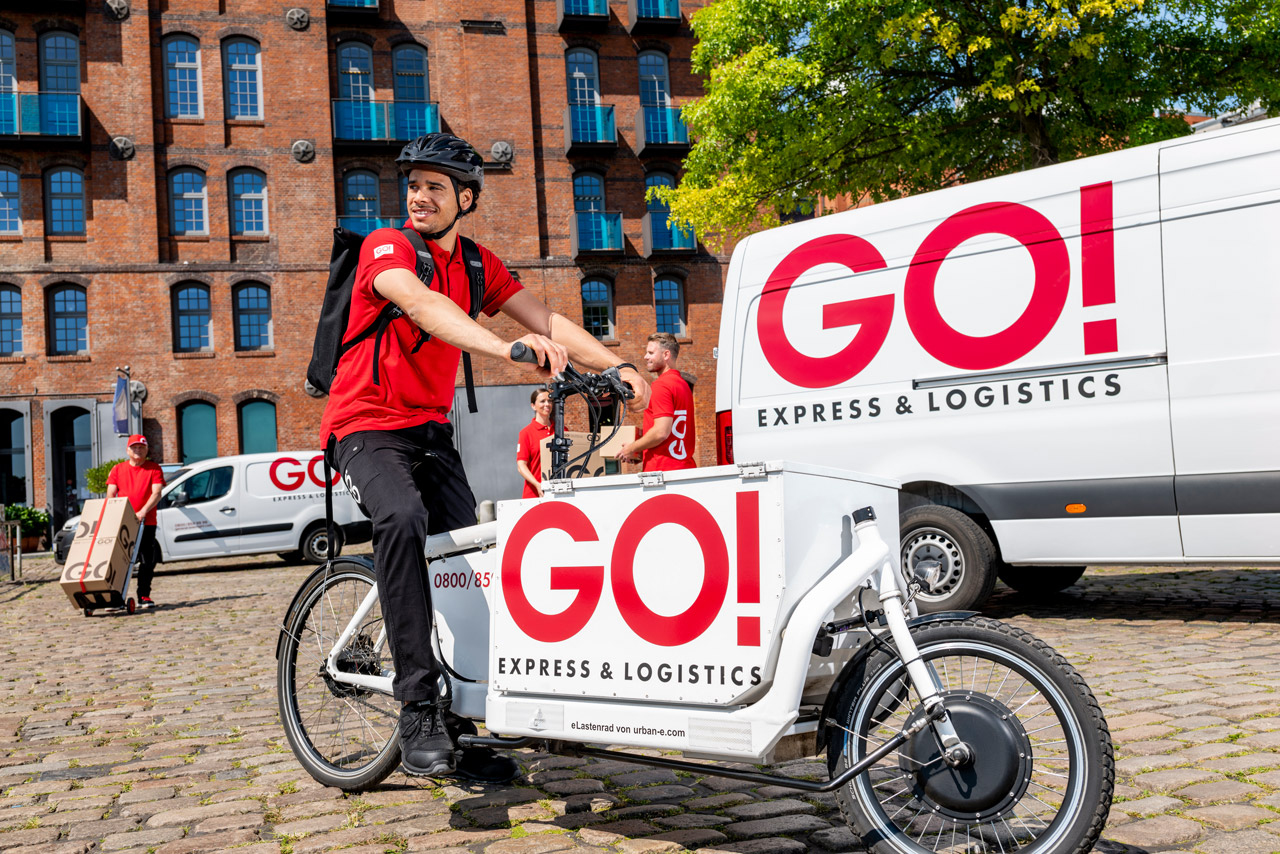 GO! Fahrradkurier auf einem Lastenfahrrad mit drei Kurieren und zwei Transportern im Hintergrund vor einem Backsteinhaus